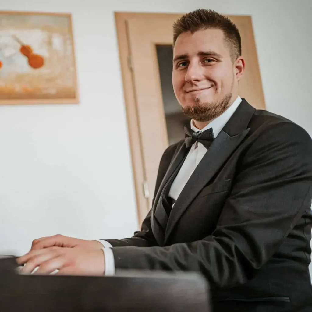 Jurica Hrenić, nasmiješen gleda u kameru, odjeven u svečano crno odijelo s leptir-mašnom, sjedi za crnim klavirom s rukama položenim na tipkama. Iza njega su bijeli zidovi ukrašeni umjetničkim slikama te meko osvijetljeni moderan interijer.