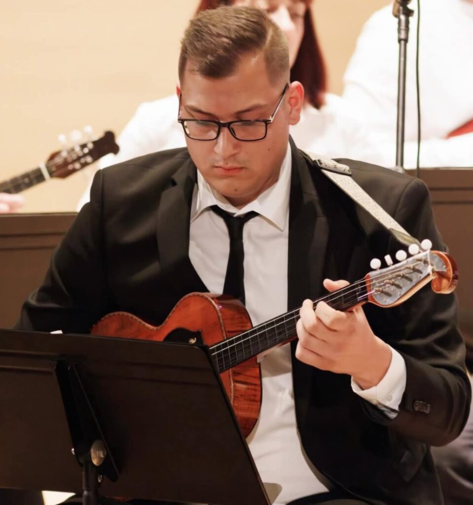 Tomislav Fajfar svira tamburaški brač u formalnom odijelu tijekom nastupa ansambla, usmjeren na notni zapis.